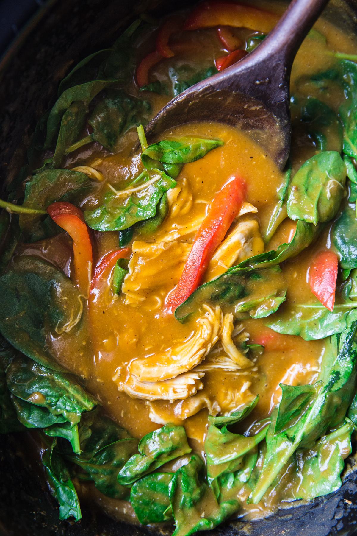 pumpkin curry with bell peppers, chicken and spinach being stirred with a wood spoon in a crock pot