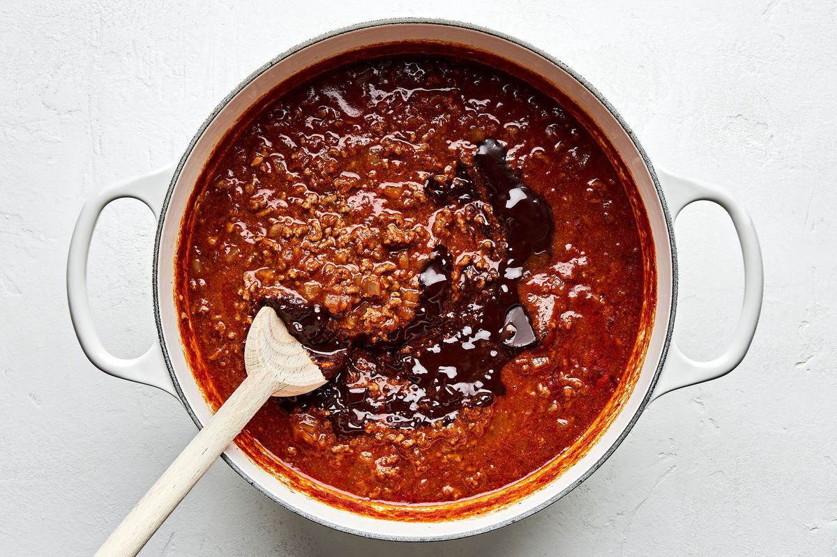 Apple cider vinegar and chocolate being stirred in to a pot of Cincinnati chili and stirred with a wooden spoon